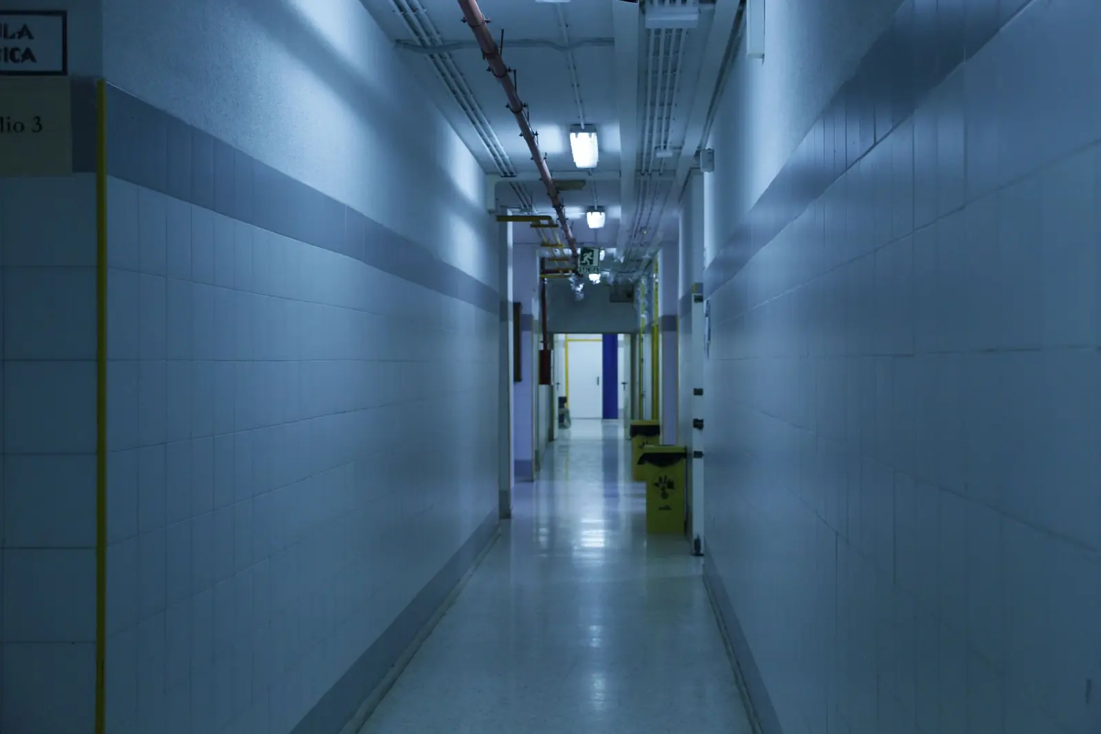 A quiet, dimly lit hospital corridor with tiled walls and overhead lights.
